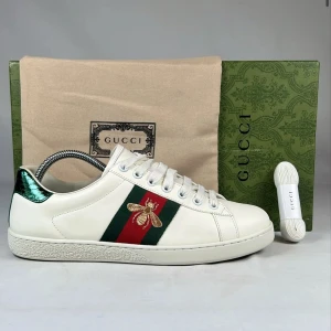 Gucci Ace sneakers med broderad bi - Säljer ett par Gucci Ace sneakers i vitt skinn med klassiska gröna och röda ränder på sidan och en broderad guldfärgad bi. Skorna har snörning, rund tå och metallic-detaljer i grönt och rött på hälen.                                                                      Märke: Gucci Modell: Ace Embroidered Bee Sneakers Färg: Vit med grön/röd Web & guldbi Skick: [Nytt/ Nästan nytt/ Lätt använt – uppdatera] Ingår: Originalbox, dustbag, extra snören Storlek: 41,5