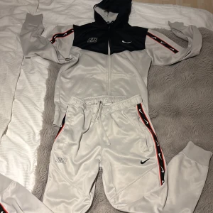 Kan röra på priset. Nike vit, svart och röd tracksuit med logoband(S) - Nike tracksuit i ljusgrått med svarta och röda logoband längs sidorna. Byxorna har snörning i midjan, muddar vid bensluten och två fickor. Hoddien är svart vid bröstet och med röd logo band med och resten ljusgrå. Materialet är mjukt och glansigt, perfekt för chill eller sport. Swoosh-logga på benet och Nike-brodyr vid fickan