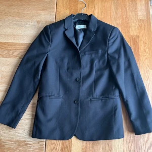 Svart kavaj från H&M - Stilren svart kavaj från H&M i storlek 146. Klassisk modell med två knappar framtill, tre dekorativa knappar vid ärmslut och tre fickor. Materialet är polyester och viskos. Perfekt för dig som gillar en enkel och snygg look. Sparsamt använd och väldigt fint skick och hel o ren.
