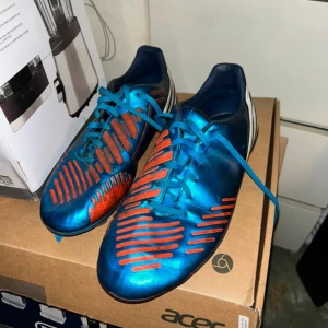 Blå och röda adidas fotbollsskor - Säljer ett par adidas fotbollsskor i metallicblå med röda detaljer och snörning. Skorna har en smal och sportig form med rund tå och platt sula, perfekta för dig som vill sticka ut på planen. Materialet är syntet och designen är riktigt modern. OBS! tvättar såklart ifall man vill köpa