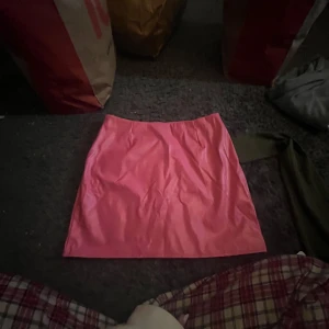 Rosa kortkjol i syntetmaterial - Säljer en snygg rosa kortkjol med slät yta och enkel design. Kjolen har en rak passform och är gjord i ett glansigt syntetmaterial som ger en cool vibe. Perfekt att matcha med en oversized hoodie eller en croppad topp för en trendig look.