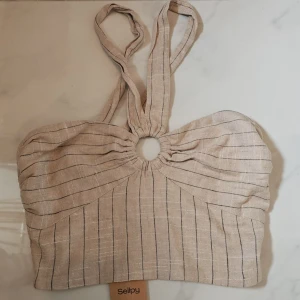 Randig halterneck topp från Zara - Snygg halterneck topp från Zara i beige-brun ton med tunna svarta ränder. Toppen har en rund ringdetalj framtill och smock på sidorna för extra komfort. Tillverkad i linne, perfekt för varma dagar och chill vibes.