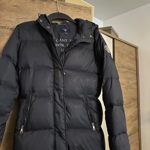 Svart dunjacka från Gant med huva - Snygg svart dunjacka från Gant med huva och två praktiska fickor med dragkedja. Jackan har en quiltad design och en patch på ärmen. Perfekt för kalla dagar och har en stilren look som funkar till det mesta.