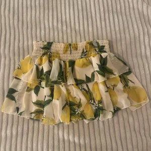 Vit kortkjol med citronmönster och volanger - Superfin kortkjol i vitt med färgglatt citronmönster och gröna blad. Kjolen har bred resår i midjan och dubbla volanger för extra flowy känsla. Tunt, luftigt tyg med innerfoder. Perfekt för sommarens alla äventyr!