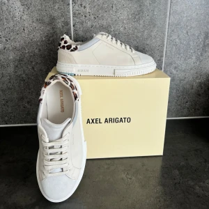 Axel Arigato vita sneakers med leoparddetalj - Snygga vita sneakers från Axel Arigato med leopardmönstrad häl och stilren design. Skorna har rund tå, snörning och platt sula. Tillverkade i skinn med mockadetaljer vid tån och hälen. Använda bokstavligen EN gång. Pris kan diskuteras.