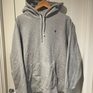 Grå Ralph Lauren hoodie - Säljer en grå hoodie från Polo Ralph Lauren med klassisk huva och snörning. Tröjan har en känguruficka framtill och den ikoniska lilla Polo-loggan broderad på bröstet. Perfekt för chill dagar och enkel att matcha med jeans eller liknande.