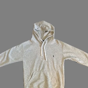 Grå hoodie från Polo Ralph Lauren - Säljer en stilren grå hoodie från Polo Ralph Lauren i storlek S. Tröjan har huva med vita snören, klassisk känguruficka och den ikoniska lilla Polo-loggan broderad på bröstet. Perfekt för chill dagar och enkel att matcha med jeans eller joggers.