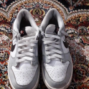 Nike Dunk sneakers i vitt och grått - Nike Dunk sneakers med vit och ljusgrå ovandel, svart swoosh på sidan och klassisk Nike-logga på hälen. Skorna har snörning, perforerad tå och platt sula. Perfekt för dig som gillar streetstyle och vill ha en clean look med retrovibe. Inkluderas crease skydd