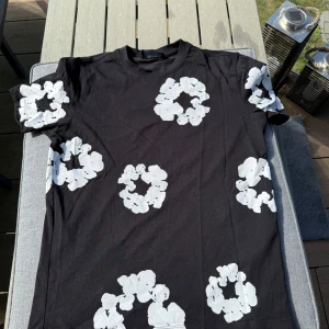 Svart Denim Tears t-shirt med blommor - Svart t-shirt från Denim Tears med vita blommiga tryck över hela plagget. Klassisk rund hals och korta ärmar. Materialet är mjuk bomull och passformen är normal. Perfekt statement-plagg med unik design för dig som gillar streetwear.