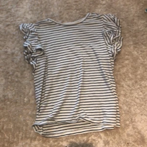 Randig blus med volangärm från POMPdeLUX - Supersöt blus i vitt med svarta ränder från POMPdeLUX. Den har korta ärmar med volangdetaljer som ger en extra touch. Mjuk och skön bomullskänsla, perfekt för dig som gillar en chill och trendig look. Rund halsringning och loose passform.