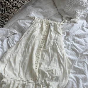 Vit linne  från H&M med spets - Supersöt vit linne från H&M i storlek 32. Klänningen har tunna axelband, omlott-design och en dekorativ spetsdetalj framtill. Materialet är mjukt och lätt bomull, perfekt för varma dagar. Passar dig som gillar romantisk och fräsch stil.