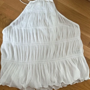 Vit plisserad linnetopp med halterneck - Superfin vit linnetopp från H&M med plisserad struktur och halterneck. Toppen har tunna band i nacken och ett flowigt, luftigt fall. Perfekt för varma dagar och ger en fräsch vibe till din outfit. Materialet är lätt och känns skönt mot huden.