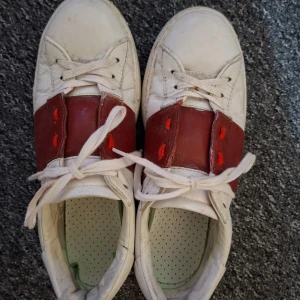 Valentino Garavani vita och röda sneakers - Snygga sneakers från Valentino Garavani med vit bas och bred röd läderpanel över mitten. Skorna har vita snören, röda detaljer vid snörningen och coola nitar runt hälen. Platt sula och rund tå ger en klassisk streetstil. Perfekta för dig som gillar lyxiga märken och unik design. De är använda så därför är priset lågt. Kan heller inte garantera äktheten på dem. Bara höhr av dig vid funderingar! 😁
