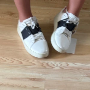 Valentino Garavani Open sneakers vit/svart - Säljer ett par Valentino Garavani Open sneakers i vitt skinn med bred svart läderdetalj över mitten. Skorna har snörning, platt sula och rund tå. Ikoniska nitar baktill ger extra edge. Perfekta för dig som gillar lyxiga och stilrena sneakers.