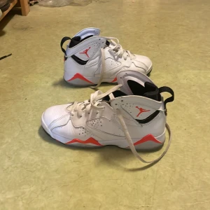 Nike Jordan 7 Retro vit/röd sneakers - Nike Jordan 7 Retro sneakers i vitt med röda och svarta detaljer. Snygg high-top modell med snörning och cool Jumpman-logga på sidan och undersidan. Skorna har en platt sula med grafiskt mönster och är tillverkade i skinn och syntetmaterial.