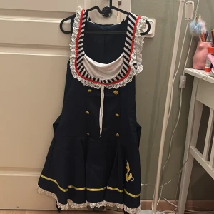 Marinblå kortklänning med ränder och spets - Säljer en marinblå kortklänning med vita och svarta ränder upptill, dekorerad med röd kant och vit spets. Klänningen har guldiga knappar, broderat ankare i gult, guldfärgade band och knyt i ryggen. Kjolen är utsvängd med spetskant. Perfekt för temafest. 