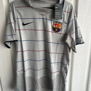 Barcelona Ronaldinho #10 fotbollströja - Snygg Barcelona fotbollströja från Nike med Ronaldinho och nummer 10 på ryggen. Tröjan är ljusgrå med tunna randiga detaljer i blått och rött, har korta ärmar och meshpaneler för extra ventilation. Klassisk klubbmärke på bröstet och Nike-logga. Perfekt för dig som älskar fotboll och retrostil.