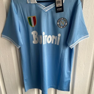Napoli Maradona 10 retro fotbollströja - Retro fotbollströja från Napoli med Maradona och nummer 10 på ryggen. Ljusblå färg med vita detaljer, klassisk krage och korta ärmar. Tryck med Buitoni på bröstet och klubbmärke samt italiensk flagga. Materialet är polyester, perfekt för fans.