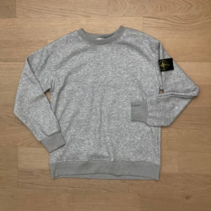 Grå sweatshirt från Stone Island - Säljer en grå sweatshirt från Stone Island med klassisk rund hals och ribbade muddar. Tröjan har den ikoniska Stone Island-patchen på vänster ärm. Mjuk insida och stilren look, perfekt för dig som gillar streetwear och vill ha något cleant i garderoben.