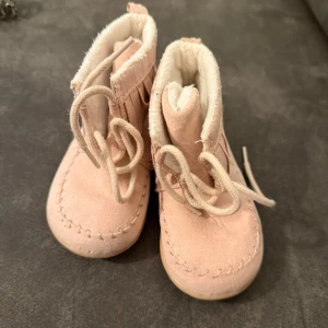 Beiga mockaboots med fransar, strl 20 - Supersöta beiga boots i mocka med snörning och dekorativa fransar runt skaftet. Insidan är fodrad med mjukt vitt tyg som gör dem extra sköna. Perfekta för små fötter som vill ha både stil och komfort.  Står ej för frakten 