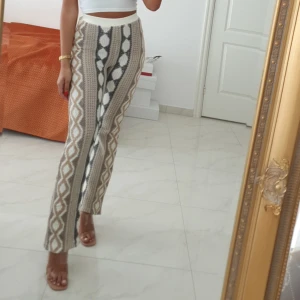 Mönstrade stickade byxor i beige och vit - Säljer ett par unika stickade byxor med högt midja och raka ben. Byxorna har ett coolt geometriskt mönster i beige, vit och svart. Perfekta för dig som vill sticka ut med din stil och gillar bekväma material.