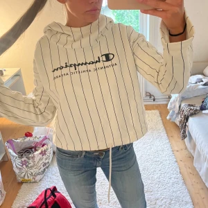 Vit randig hoodie från Champion - Säljer en vit hoodie från Champion med svarta vertikala ränder och logga på bröstet. Tröjan har huva, långa ärmar och snörning nedtill. Perfekt för dig som gillar sportig och chill stil. Passar dig som vill ha en loose fit.