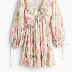 Blommig kortklänning med volang - Superfin kortklänning med plisserat tyg och blommigt mönster i ljusa toner av rosa, grönt och beige. Klänningen har v-ringad hals, volangdetalj framtill och långa ärmar med snörning vid ärmslut. Perfekt för sommarens alla tillfällen!