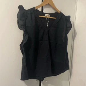 Svart blus med volang från Sunflower - Svart blus från Sunflower med volangdetaljer på axlarna och v-ringad hals. Blusen är ärmlös och har en luftig, loose passform som ger en chill vibe. Perfekt för dig som gillar stilrena plagg med lite extra edge. Materialet känns lätt och bekvämt mot huden.