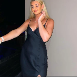 Svart omlottklänning med smala axelband - Supersnygg svart långklänning med omlott och v-ringad hals. Klänningen har smala axelband och dekorativa knappar längs sidan. Materialet är glansigt och känns lyxigt, perfekt för att sticka ut. Passformen är normal och klänningen har en liten slits framtill.