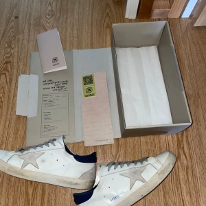 Golden Goose vita sneakers med stjärna - Golden Goose sneakers i vitt skinn med grå snörning och en ikonisk grå stjärna på sidan. Skorna har mörkblå detaljer vid hälen och en platt brun sula med fiskbensmönster. Perfekt för dig som gillar en cool och avslappnad stil. Priset kan diskuteras.