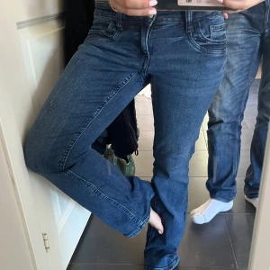 Blå bootcut jeans med låg midja - Snygga blå jeans med bootcut passform och låg midja. Klassisk femficksmodell med dragkedja och knapp framtill. Jeansen har en mörkblå tvätt och är gjorda i jeansmaterial med lite stretch för skönare passform. Perfekta för dig som gillar retrovibbar och vill ha ett par jeans som sticker ut. Storlek 158