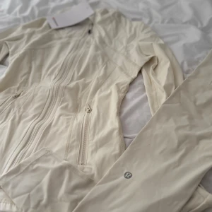Beige vindjacka från Lululemon - Snygg beige vindjacka från Lululemon med figurnära passform och långa ärmar. Jackan har flera dragkedjor framtill och praktiska fickor. Tillverkad i lätt syntetmaterial som är perfekt för träning och aktiva dagar. Diskret Lululemon-logga på ärmen.