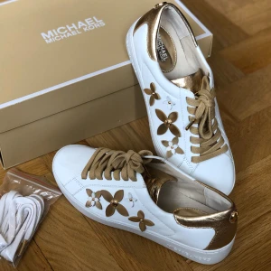 Michael Kors vita sneakers - Michael Kors sneakers – nya, strl 36 Helt nya och oanvända sneakers i vitt läder med lyxiga gulddetaljer. Guldfärgad hälkappa, plös och dekorativa blommor i guld på sidorna. Kommer med extra vita skosnören. Perfekta för både vardag och fest, bekväma och stilrena.