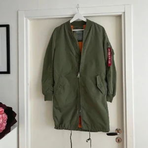 Grön lång bomberjacka Alpha Industries - Säljer en grön lång bomberjacka från Alpha Industries med orange foder och klassisk röd tag på ärmen. Jackan har snörning nedtill, dragkedja framtill och två stora fickor med lock. Perfekt för dig som gillar streetwear och utility vibes.