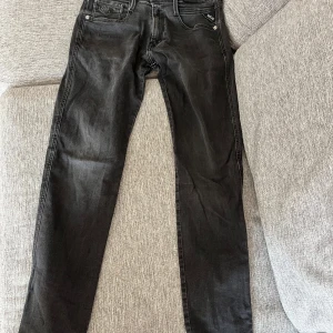 Replay Anbass svarta jeans W29 L32 - Svarta Replay Anbass jeans med klassisk femficksdesign och snygga detaljer på bakfickan. Slimmad passform och raka ben, tillverkade i mjukt jeansmaterial med dragkedja i gylfen. Perfekta för dig som gillar stilrena och moderna jeans.