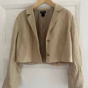 Beige croppad kavaj från Lindex - Snygg croppad kavaj från Lindex i beige linne. Klassisk krage, två knappar framtill och långärmad modell. Perfekt för dig som gillar stilrena och trendiga plagg med en modern twist. Passar dig som vill ha en lätt och luftig jacka med cool siluett.