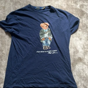 Marinblå Polo Bear t-shirt Ralph Lauren - Snygg marinblå t-shirt från Polo Ralph Lauren med klassisk Polo Bear-tryck på bröstet. T-shirten har rund hals och korta ärmar, tillverkad i mjuk bomull.