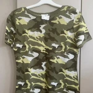 Camo topp med kort ärm - Cool camouflagemönstrad topp i grönt, beige och vitt. Modellen har rund halsringning och korta ärmar. Materialet är mjukt och stretchigt med 70% viskos och 30% polyamid, vilket gör den skön att bära. Perfekt för dig som gillar streetstyle och vill sticka ut.