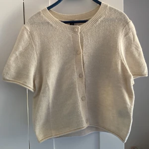 Beige kortärmad kofta från H&M - Säljer en beige, kortärmad kofta från H&M med rund hals och knappar framtill. Koftan är stickad i mjukt material och har ribbade detaljer vid hals och knappslå. Perfekt för lager-på-lager och chill stil.