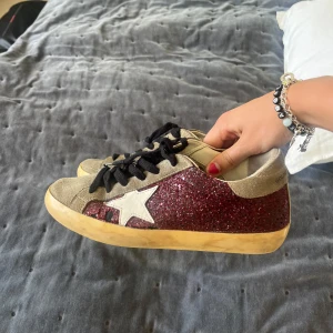 Golden Goose glitter sneakers - Säljer ett par Golden Goose sneakers med vinröda glittriga sidor, beige mockadetaljer och vit stjärna på sidan. Skorna har svarta snören och en platt, gulnad sula (det är meningen). Så coola! 
