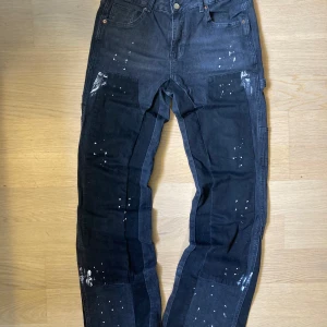 Svarta cargopants med färgstänk - Unika svarta cargopants med coola vita färgstänk och patchade detaljer på benen. Byxorna har flera fickor både fram och bak, samt råa kanter vid bensluten för en edgy vibe. Perfekt för dig som gillar streetstyle och vill sticka ut.