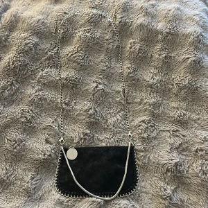 Svart axelväska med kedja - Snygg svart axelväska med silverfärgad kedja och rund bricka. Väskan har en mjuk mockaliknande yta och dekorativa kedjedetaljer runt kanterna. Stängs med dragkedja och har ett rymligt fack inuti. Perfekt för att lyfta din outfit med lite edge.