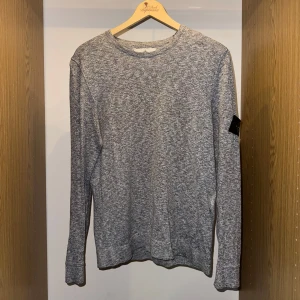 Grå långärmad tröja Stone Island - Snygg gråmelerad långärmad tröja från Stone Island med klassisk rund hals och ikonisk patch på vänster ärm. Tröjan har en normal passform och är tillverkad i mjuk bomull som är perfekt för chill dagar. En stilren basic med street-vibe.