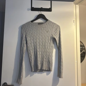 Grå kabelstickad tröja från GANT - Säljer en snygg grå kabelstickad tröja från GANT i stretchig bomull. Tröjan har rund hals, lång ärm och GANT-logga broderad på bröstet. Perfekt för dig som gillar klassisk stil med en modern twist. Passar dig som vill ha en slim passform.