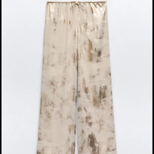 Beige och guldiga wide byxor med snörning - Säljer ett par beige byxor med guldiga detaljer och mönster. De har en bred, loose passform och snörning i midjan för extra komfort. Materialet är glansigt och känns lyxigt, perfekt för dig som gillar att sticka ut med coola detaljer.