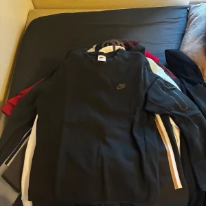 Svart Nike sweatshirt med logga - Svart sweatshirt från Nike med diskret logga på bröstet. Tröjan har rund hals och långa ärmar, samt en ficka med dragkedja på ena ärmen. Perfekt för dig som gillar enkel och sportig stil. Materialet känns mjukt och är troligtvis bomull eller polyester.