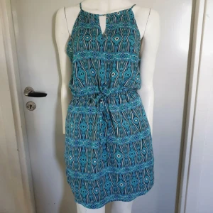 Turkos mönstrad kortklänning med snörning - Snygg kortklänning i turkos och svart med grafiskt mönster. Klänningen har smala axelband, nyckelhålsöppning fram och knyt i midjan för en skön passform. Tillverkad i lätt och luftig polyester med ett tunt innerfoder. 