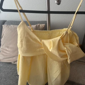 Gul omlott blus med volangdetalj - Supersöt ljusgul blus med omlott och volangdetaljer. Toppen har en axelbandslös design på ena sidan och smala axelband på andra, vilket ger en cool asymmetrisk look. Materialet är lätt och krispigt, perfekt för varma dagar.