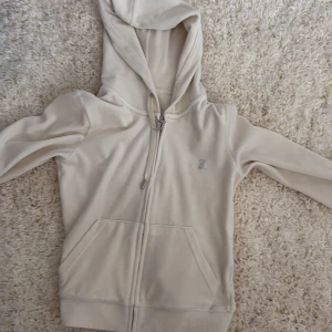 Vit Juicy Couture hoodie med strass - Säljer en vit hoodie från Juicy Couture i mjuk sammet med strassdetaljer på ryggen där det står 'Juicy'. Tröjan har huva, dragkedja framtill och två fickor. Kontakta vid frågor!