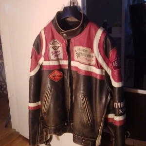 Arturo Motors svart och röd skinnjacka - Säljer en riktigt cool svart och röd skinnjacka från Arturo Motors med vita detaljer och broderade patches. Jackan har ståkrage, dragkedja framtill, två fickor med dragkedja och snygga racing-inspirerade emblem. Perfekt för dig som gillar bikerstil och vill sticka ut.
